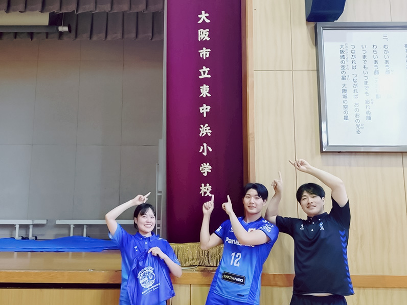 写真：学校名の前でポーズをとる伊藤マネージャー、竹内マネージャー、伊藤アカデミーコーチ