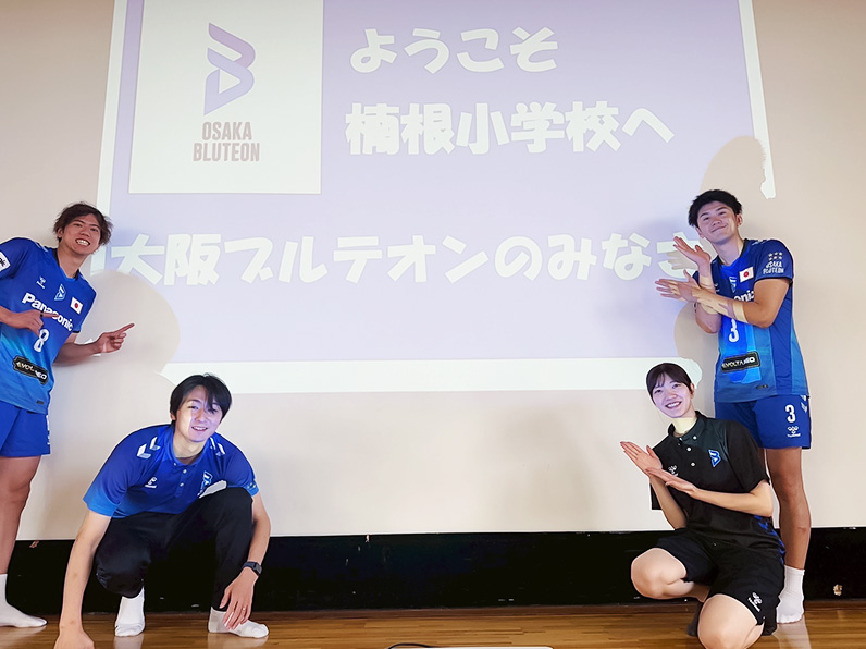写真：学校名の前でポーズをとる仲本選手、牧山マネージャー、西條アカデミーコーチ、中村選手